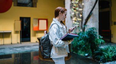 Barcelona 'daki eski bir binanın avlusunda dururken elinde defterler ve akıllı telefonları tutan genç bir kadının yan görüntüsü.