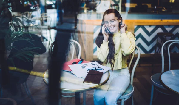 流行のメガネと明るい黄色のトレーナーの魅力的な若い女性は 電話で話しながら髪を調整し 居心地の良いモダンなカフェに座って — ストック写真
