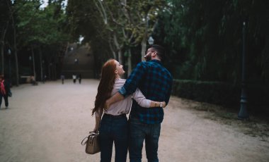 Gündüz vakti kucaklaşan ve parkta yürürken birbirlerine bakıp gülümseyen neşeli çiftin arka planı.