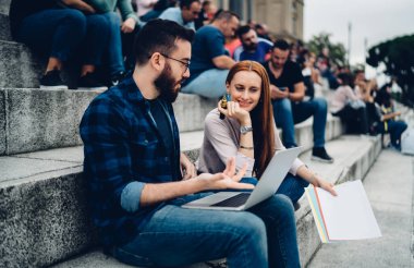 Genç sakallı erkek, gün ışığında kalabalık merdivenlerde otururken kızıl saçlı kız arkadaşına üniversite ödevinde yardım ediyor.