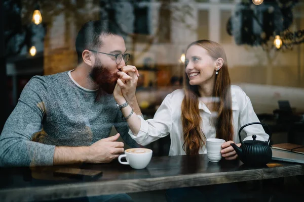 在现代咖啡馆里 一个可爱的年轻人坐在窗后 彼此凝视 亲吻着女人的手 — 图库照片