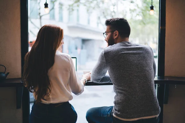 Back view of content youthful couple sitting at small table in cozy cafe near big window and looking at each other while drinking coffee