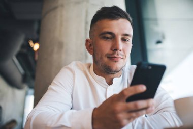 Gündüz vakti sokak kafesinde dinlenirken akıllı telefon kullanan beyaz tişörtlü kahverengi saçlı genç adama odaklanmış.