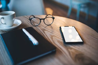 Kahve fincanının yanında beyaz kalem ve gözlüklü siyah defter ve kafeteryadaki ahşap masada boş ekranlı akıllı telefon.