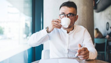 Beyaz gömlek ve gözlük takan serbest çalışan bir erkek. Yaratıcı kafeteryadaki uzak çalışma sırasında günlük planlamada not alırken bir yandan da kahve içiyor.