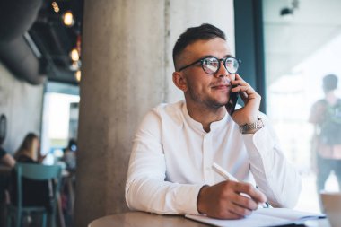 Yaratıcı kafeteryada çalışırken, telefon görüşmeleri sırasında beyaz gömlek ve gözlük takan, notlar yazan serbest çalışan genç bir erkek.