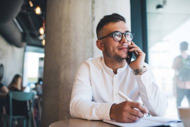 Focused young male freelancer wearing white shirt and eyeglasses writing notes during phone call while working remotely in creative cafeteria clipart
