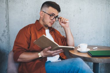 Gözlüklü, kırmızı tişörtlü, kot pantolonlu, düşünceli, gözlüklü, günlük planlamacıdan notlar alan ve elinde kahve fincanıyla yaratıcı kafenin gri beton duvarının yanında oturan genç ve düşünceli bir adam.