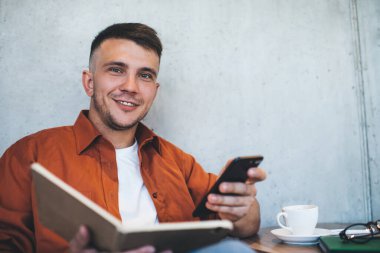 Kahve masasında oturup kameraya bakarken cep telefonuyla mesajlaşan ve notlar okuyan neşeli genç bir erkek.