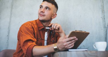 Turuncu gömlekli ve kol saati takan düşünceli genç adam kafeteryada elinde gözlüklerle oturuyor ve gözlerini kaçırıyor.