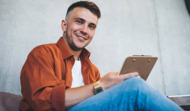 Tıraşsız, renkli gömlekli, kameraya gülümseyen ve iş sırasında sandalyede otururken elinde pano tutan pozitif bir genç.