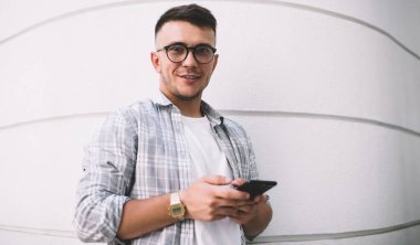 Pozitif kareli gömlek ve tişört giyen, aksesuarlı, cep telefonuna bakıp dişlek gülümseyen adam.