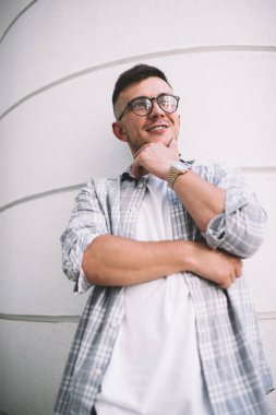 Pozitif, gözlüklü, çeneye dokunan, beyaz bir binaya çizgilerle bakan genç bir adam.