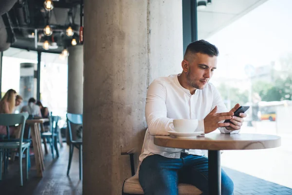 Konsentrert Mannlig Frilanser Som Leser Internett Mobiltelefon Drikker Kaffe Mens – stockfoto