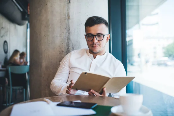Young calm male freelancer wearing white shirt and glasses reading daily organizer while sitting at table with smartphone and cup of coffee near window during remote work in creative cafeteria