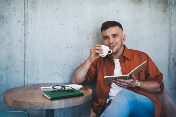 Positive young man in casual clothing enjoying cup of coffee while chilling with organizer in comfortable youth cafe at daytime