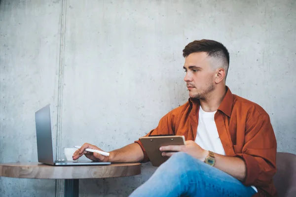 Pensive young male with beard and short haircut in casual clothes sitting near table and browsing laptop while working on project
