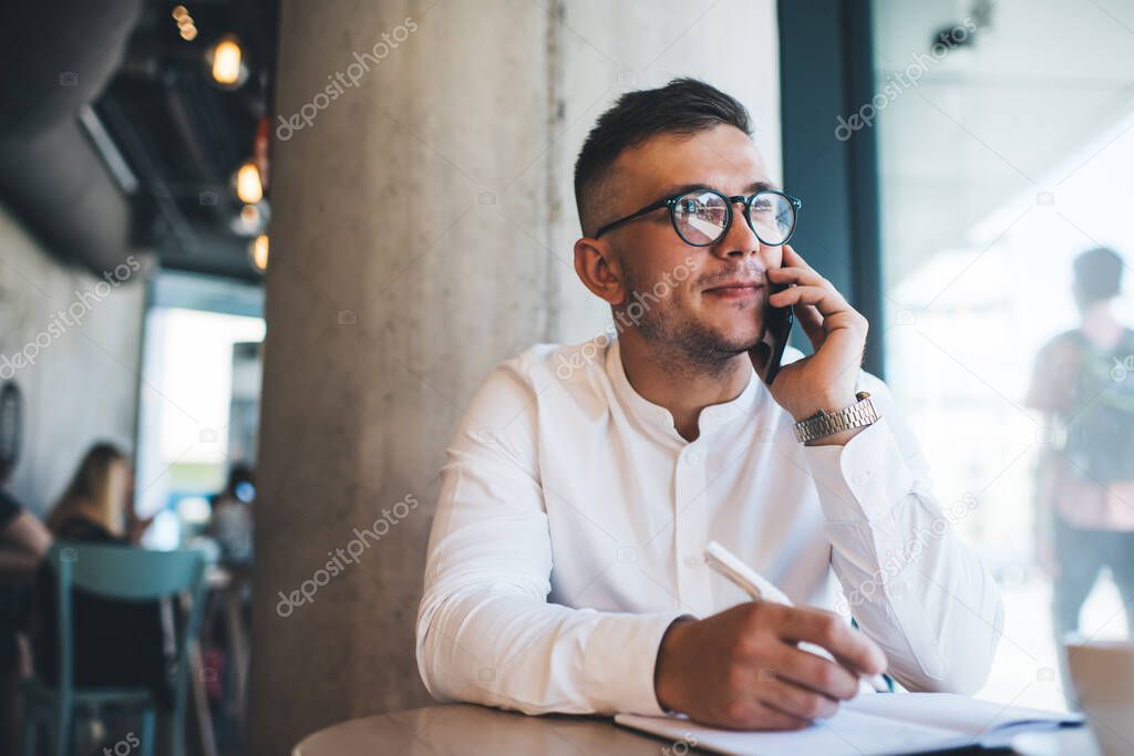 Focused young male freelancer wearing white shirt and eyeglasses writing notes during phone call while working remotely in creative cafeteria