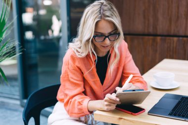 Kendine güveni tam bir serbest çalışan, gözlük takan, dizüstü bilgisayar, akıllı telefon ve kafeteryada çalışırken kahve içerek not alan bir kadın.