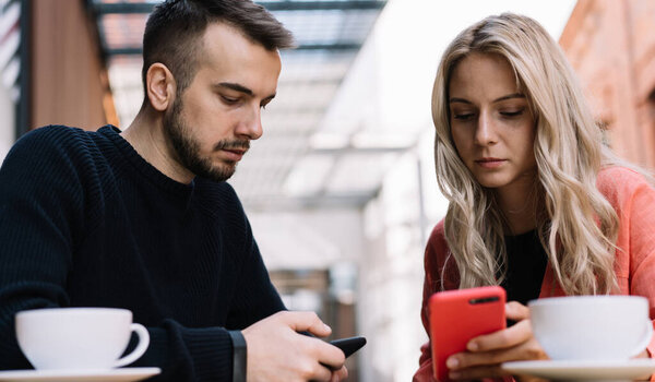 Сконцентрированная на экране молодая пара в повседневной одежде просматривает смартфоны и пьет кофе на террасе кафе в солнечный день