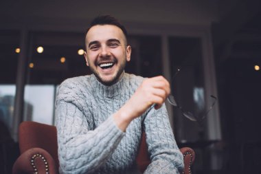 Sıradan giyinen, rahat bir kafede eğlenirken gülümseyen ve kameraya bakan düşük açılı sakallı adam.