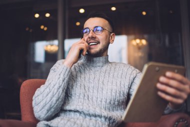 Sıradan kıyafetler ve gözlüklerle gezen, elinde bir not defteri tutarken gülümseyen ve başka tarafa bakan, serbest sakallı bir erkek.