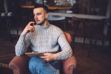 Düşünceli, sakallı, sıradan kıyafetleri olan bir erkek. Kafede elinde kalem ve not defteriyle oturuyor.