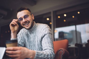 Pozitif genç adam yuvarlak gözlüklerin tapınağına dokunuyor ve rahat bir odada otururken parlak bir gülümsemeyle kameraya bakıyor.