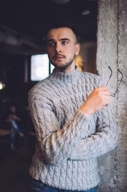 Ciddi sakallı, sıcak örülü kazaklı genç bir erkek gri duvara yaslanmış, elinde gözlüklerle karanlık odada dikkatlice başka yere bakıyor.