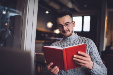Sıcak süveter ve gözlüklü olumlu sakallı bir erkek kafede oturuyor ve tatilde işten dinlenirken kitap okuyor.