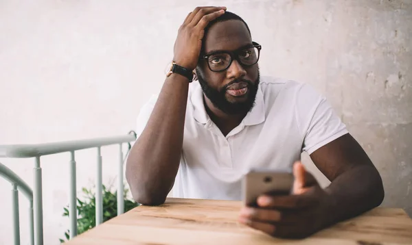 Smart African American entrepreneur in casual t shirt and eyeglasses looking at camera while solving problems with clients on mobile phone in modern cafe