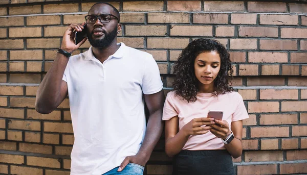 Serious dark skinned male talking on mobile phone while his girlfriend using mobile phone for chatting in social networks, african american millennial 20s woman and man concentrated on gadgets