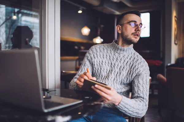 穿着休闲装和眼镜的认真的男性创业者 一边坐在桌旁 一边用笔记本电脑 一边回头看 一边在计划者中写下商业策略 — 图库照片