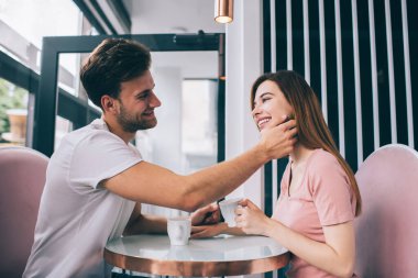Kahve için flört eden ve birbirlerine gülümseyen mutlu, duygusal çiftin yan görüntüsü. Bu arada erkek arkadaşı da nazik ve sevilen bir kadına dokunuyor.