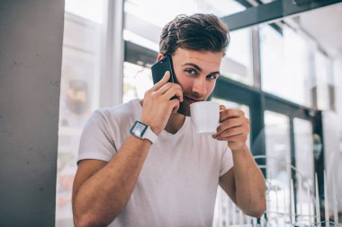Modern kafede kahve içerken kameraya bakıp cep telefonuyla konuşan, tişörtlü, şık, yakışıklı, tatmin olmuş genç bir erkek.