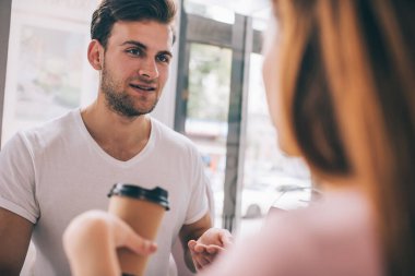 Beyaz tişörtlü sakallı genç adam kafede kahve içen bir kadınla konuşuyor.
