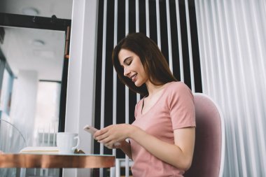 Uzun siyah saçlı çekici bir kadının akıllı telefonuyla arkadaşlarıyla sohbet etmesi modern kafede bir fincan kahve ve kitapla masada oturması.