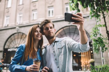 Sakin yetişkin bir adam ve çekici bir kadın sıcak bir şeyler içmeye gidiyorlar. Akıllı telefonuyla selfie çekiyorlar. Gündüz vakti eski binanın bulanık yüzüne karşı sokakta duruyorlar.