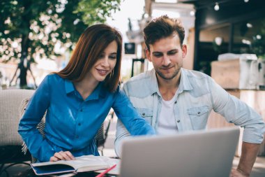 Gündelik modern adam ve dizüstü bilgisayarlı akıllı kadın sokak terasında dizüstü bilgisayarda oturuyor ve gün ışığında cihazları paylaşıyor.