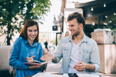 Gündelik giysiler içinde gülümseyen, eğlenceli bir eşle ödev yapan ve yaz kafeteryasında gün ışığında ders kitabını işaret eden mutlu bir erkek.