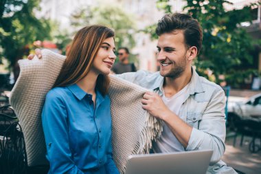 Mutlu adam, yeşil ağaçların karşısındaki kafeteryada dizüstü bilgisayarı olan sıcak ekoseyle sarmalayan mutlu kadın.