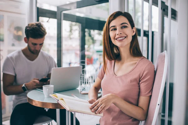 Happy modern female entrepreneur with open diary in hands smiling and looking away while sitting in cafe with male colleague