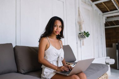 Yumuşak koltukta oturan ve Bali 'de vakit geçirirken bilgisayarla internetten çalışan yaz kıyafetli hoş bir genç bayan.