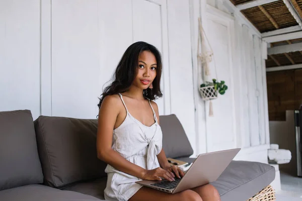 夏の衣装を着た魅力的な若い民族の女性は 柔らかいソファに座って バリで時間を過ごしながら自宅からラップトップでオンラインで働いています — ストック写真