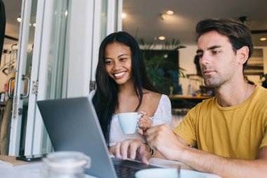 Dizüstü bilgisayara odaklanmış genç bir adam bir yandan bir kadınla çalışırken bir yandan da kahve içip modern kafede gülümsüyor.