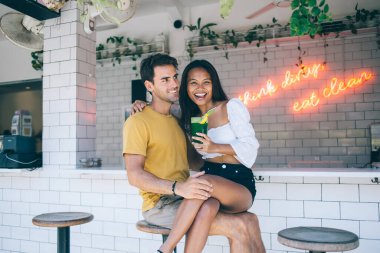 Ellerinde kokteyl olan çekici bir kadın tatmin olmuş erkek arkadaşının kucağında oturuyor ve flört ederken gülüyor ve plaj barında sarılıyor.