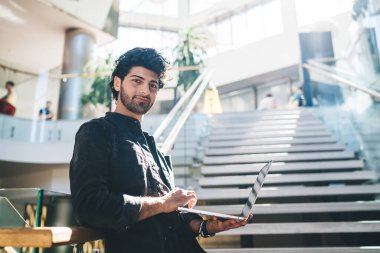 Orta Doğulu girişimcinin yarım boy portresi modern dizüstü bilgisayarı elinde tutuyor ve kameraya bakıyor. Dijital netbook ile içeriden poz veriyor.