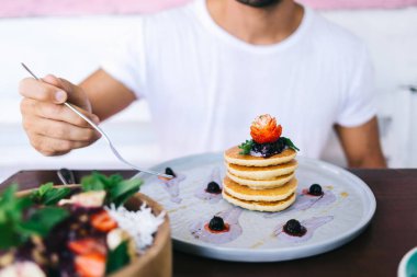 Kırpılmış görüntüde erkek, yemek masasında oturmuş sağlıklı beslenme için yemek yiyor, soslu gözleme ve çilekli meyve ve tatlı olarak düşük kalorili meyve salatası ile kase yiyor.