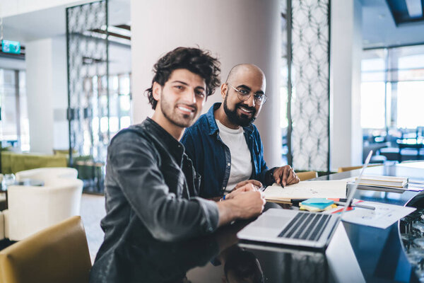 Portrait of cheerful male professionals smiling at camera during collaborative meeting for developing business project, Middle Eastern executive managers using laptop computer and textbook documents