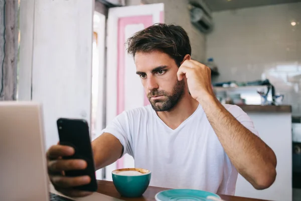 Homem Barbudo Jovem Pensativo Roupas Casuais Usando Smartphone Enquanto Bebe — Fotografia de Stock